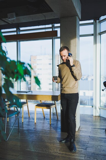 Positive young man smiling and talking on mobile phone and drinking coffee while resting while standing in office Lunch break during work