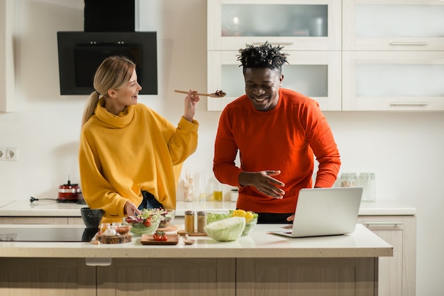 Positive young man standing in the kitchen in front of modern laptop and smiling while his happy girlfriend offering him a spoonful of salad