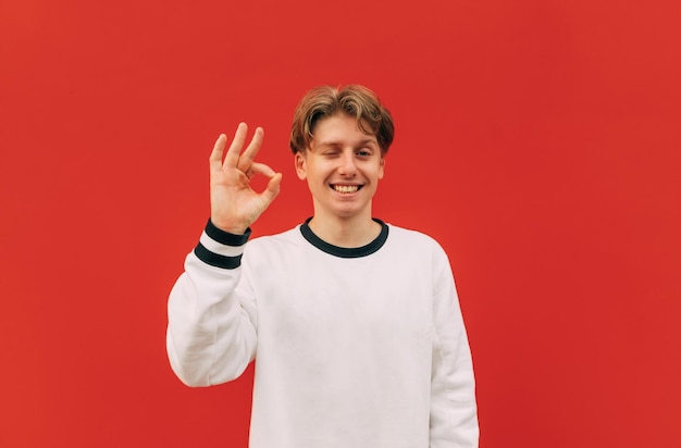Positive young man in a white sweatshirt stands on a background of a red wall looks at the camera