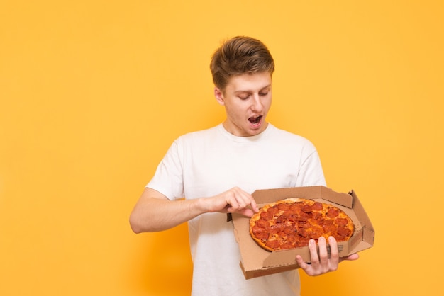 Positive young man in a white T-shirt stands on a yellow background and looks surprised at the box