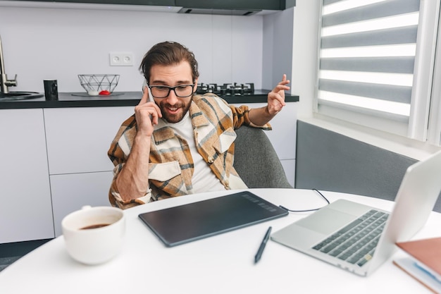 写真 彼の机に座って携帯電話で話しているひげを持つ肯定的な若い男