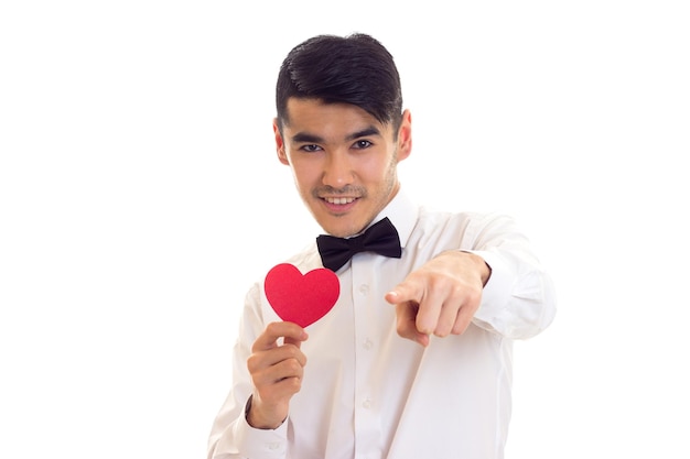 Позитивный молодой человек с темными волосами в белой футболке с черным галстуком-бабочкой держит красное бумажное сердце на белом фоне в студии