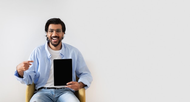 Positive young middle eastern male in glasses sits on armchair points finger at tablet with empty
