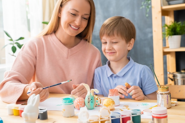 Positive young mother sitting at table and making rabbit design for Easter eggs with her son