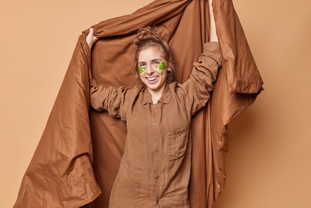 Positive young pretty woman dressed in pajama applies green\
hydrogel patches to remove puffiness and wrinkles holds soft\
blanket smiles gladfully isolated over brown background awakening\
concept