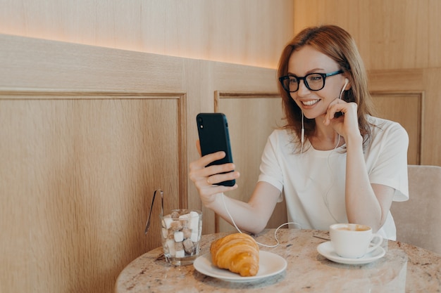 긍정적인 젊은 빨간 머리 여성 블로거는 최신 휴대전화를 들고 추종자들과 화상 채팅을 합니다.