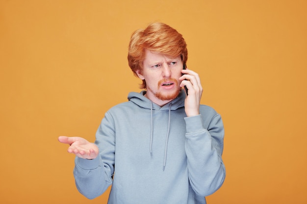 Positive young redhead man with beard standing against orange wall and pointing up while telling about promotion