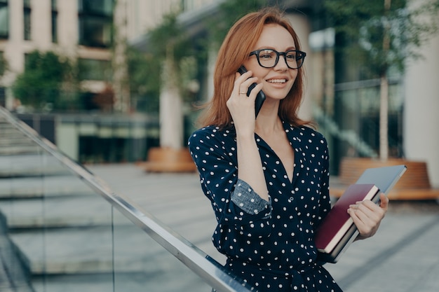 ポジティブな若い赤毛の女性は携帯電話を楽しんでいますスマートフォンを耳の近くに保ちます