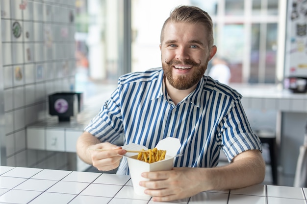 Позитивный молодой стильный парень ест китайскую лапшу в кафе во время перерыва на работе. Концепция чего-либо