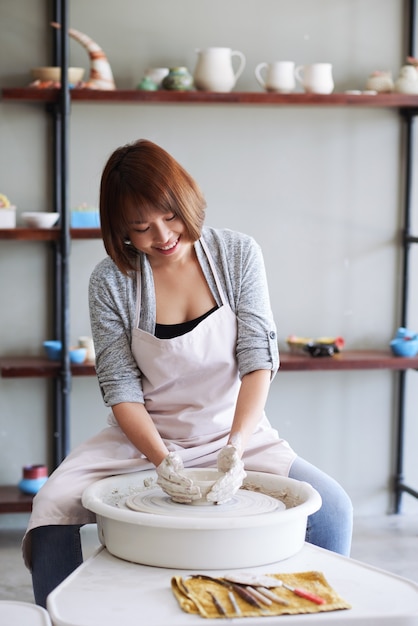 Positive young vietnamese female ceramist making clay product on potters wheel in studio