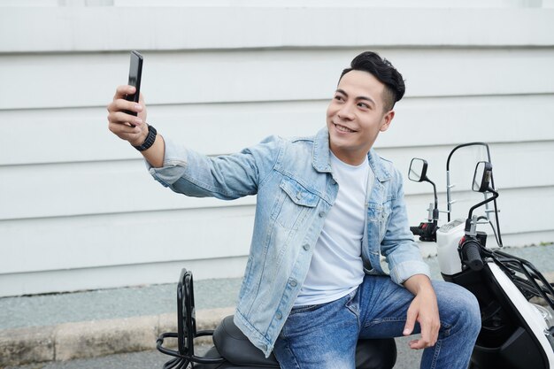 Positive young Vietnamese man in denim clothes sitting on moped and taking selfie on smartphone against street building