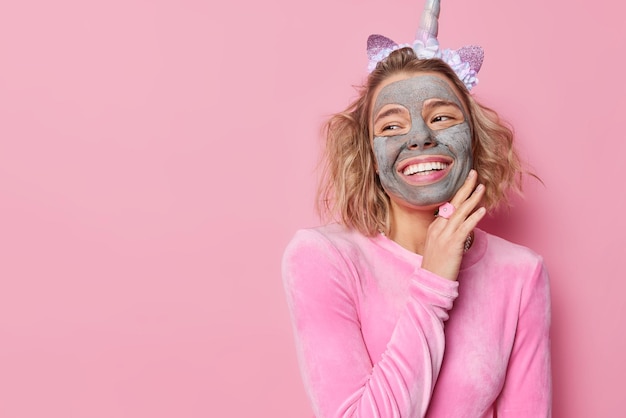 Positive young woman applies nourishing clay mask for skin treatment touches jawline gently smiles broadly wears festive outfit isolated over pink background with blank space for your promotion