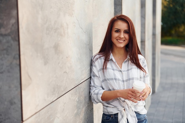 Positive young woman in casual clothes standing outdoors near wall with cup of drink in hands