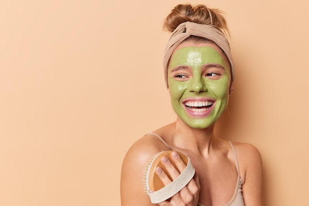 Positive young woman feels glad uses dry brush for body treatment undergoes wellness procedures applies nourishing green mask on face stands sideways with bare shoulders indoor against beige wall