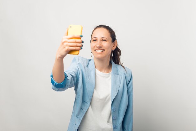 Positive young woman is taking a selfie in a studio over white background.