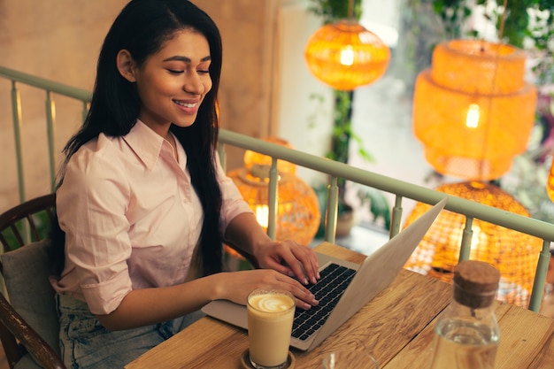 Positive young woman in jeans and a blouse sitting in a comfortable cafe alone and smiling while using her modern laptop