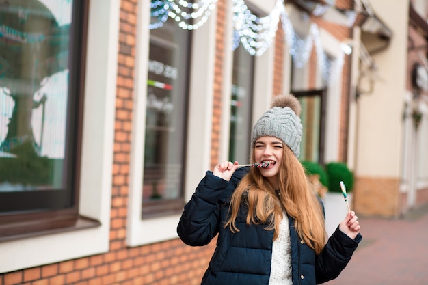 ぼやけた背景でカラフルなクリスマスキャンディーを食べるニットの暖かい帽子の肯定的な若い女性