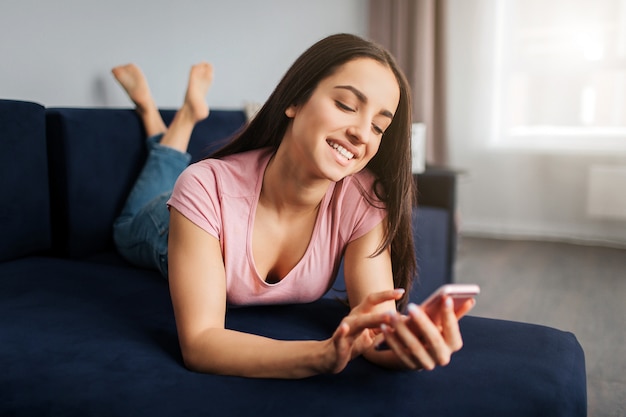 Положительная молодая женщина лежа на софе в комнате. Она касается экрана телефона и улыбается. Модель отдыхает.