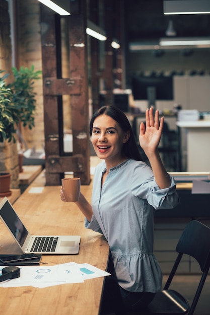 Positive young woman sitting in front of a laptop with a mug
and waving her hand