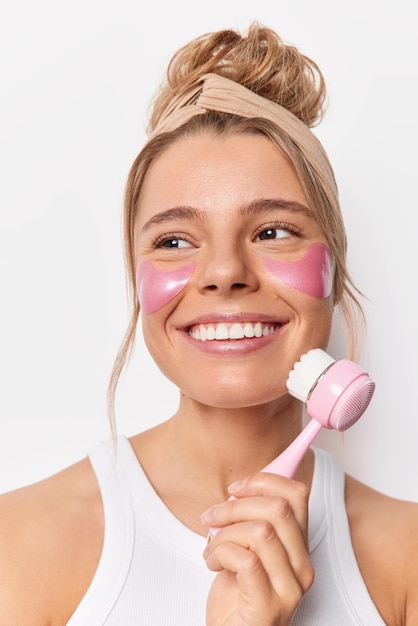 Positive young woman wears headband applies pink hydrogel patches under eyes to reduce puffiness uses face massager wears casual t shirt isolated over white background. Beauty procedures at home