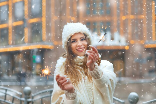 Positive young woman wears white knitted hat and coat having fun with sparklers during the snowfall