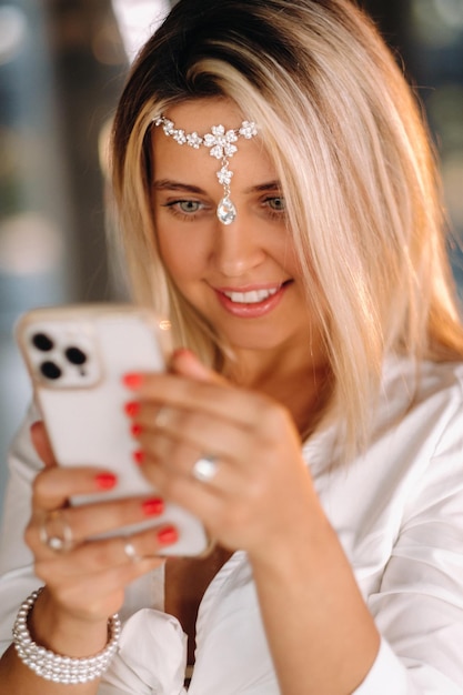 A positive young woman in a white dress and an ornament on her head was smiling holding a phone in her hands and looking into it