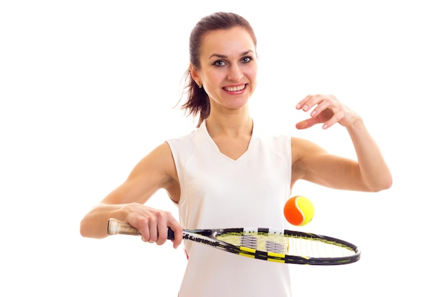 Positive young woman in white sports shirt with dark ponytail holding tennis raquet and orange ball