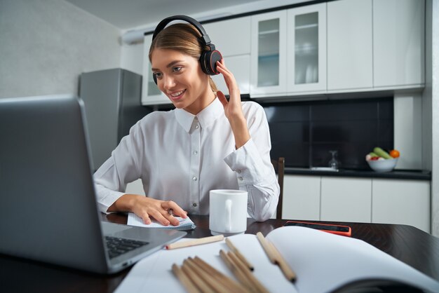 Positive young woman in wireless headphones having video chat on laptop while sitting at table