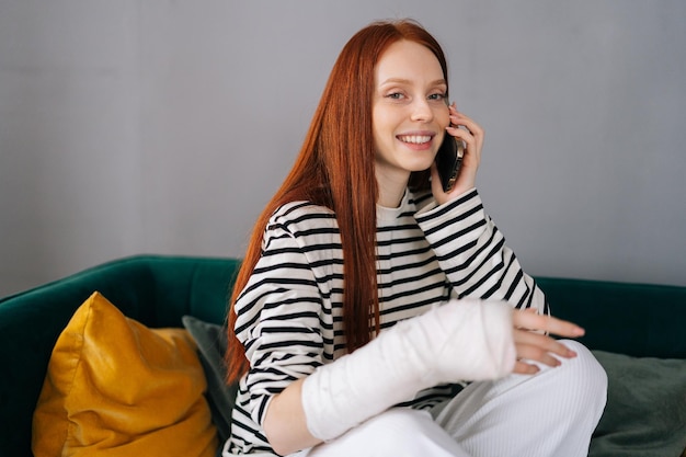 Positive young woman with broken arm wrapped in white gypsum bandage talking on smartphone with friends during rehabilitation looking at camera