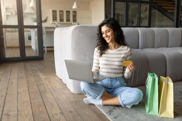 Positive young woman with gift bags using laptop and credit card for online shopping in web store