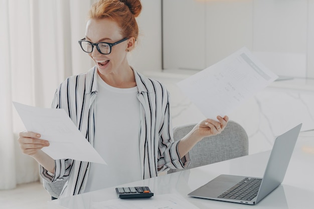 Positive young woman with red hair calculates utility charges engaged in planning monthly budget