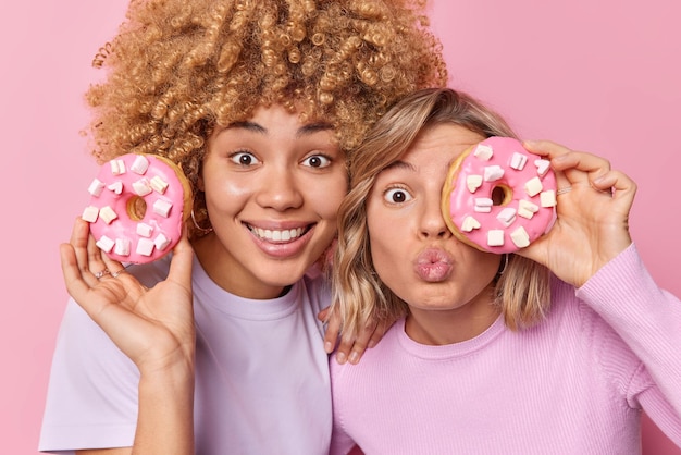 Positive young women hold delicious doughnuts with marshmallow eat tasty food stand next to each other dressed casually isolated over pink background Unhealthy nutrition and binge eating concept