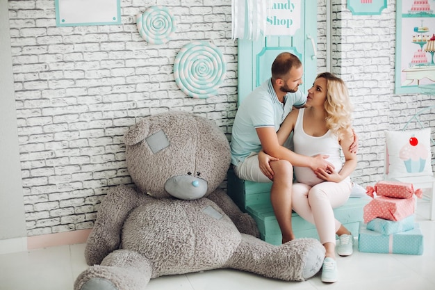 Positivity couple of handsome man and pretty blonde pragnant woman sitting on stairs at studio embracing stomach and gesturing heart by hands Young stylish family at sweet mint interior among toys