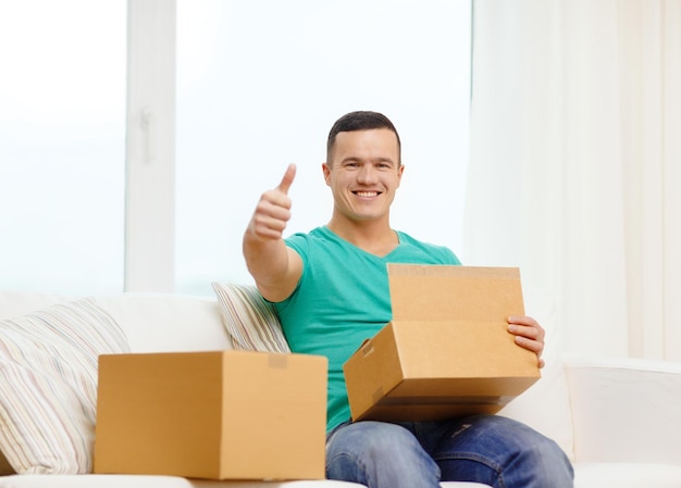 почта, дом и концепция образа жизни - улыбающийся мужчина с картонными коробками дома показывает палец вверх