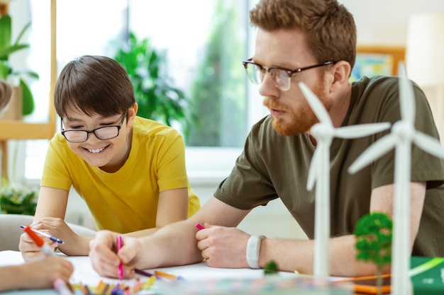 Poster about environment. Smiling dark-haired boy being interested in cognitive project while drawing next to his teacher