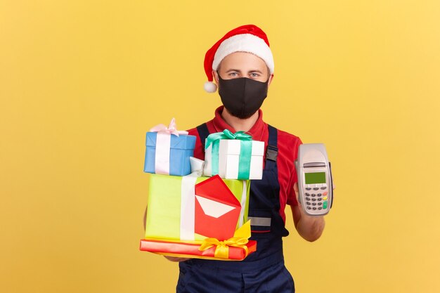 Postman in santa claus hat with medical protective mask holding gift boxes and payment terminal