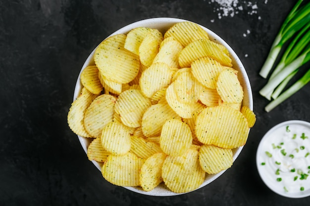 Potato chips Crispy potato chips and sour cream with onion in bowl on a dark stone concrete background top view with copy space