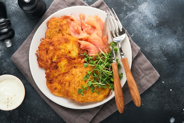 Potato pancakes Fried homemade potato pancakes or latkes with cream green onions microgreens red salmon and sauce in rustic plate on old black table background Rustic style Top view