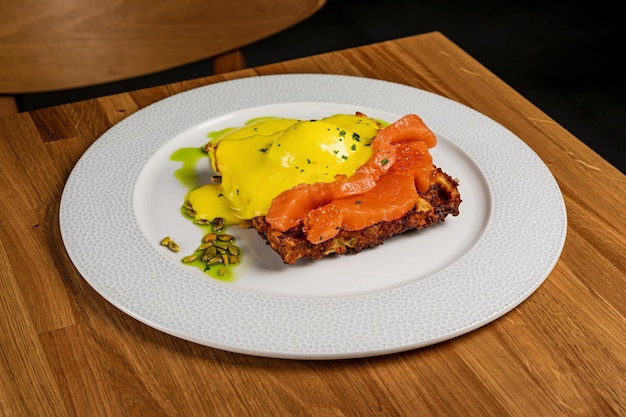 A potato waffle with salmon poached egg caviar and bearnaise sauce served on a white plate on a wooden table of a restaurant