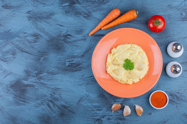 Purea di patate su un piatto accanto a ciotole di verdure e spezie, sul blu.