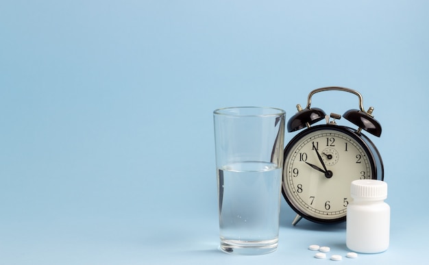 Potje pillen, water en een klok op een blauwe tafel. Tijd om pillen te slikken. Slapeloosheid. Kopieer ruimte.