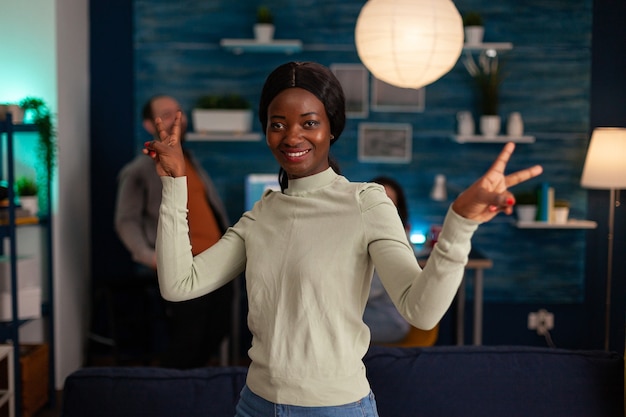 Портрет афро-американской женщины, улыбающейся в камеру, показывает знак рукой победы поздно ночью в гостиной. На заднем плане многонациональные друзья собираются вместе и веселятся во время вечеринки по выходным.