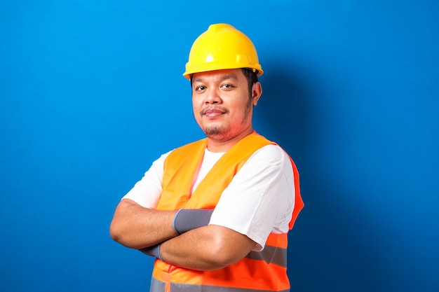 Potrait of fat asian worker wearing helmet and safety vest looking to camera crossed his hand with confidence gesture against blue background