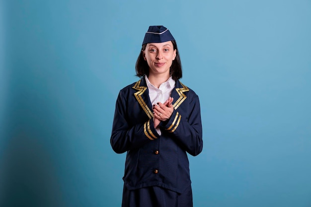 Potrait of smiling flight attendant clapping hands while standing in studio with blue background. Stewardess wearing aviation uniform applauding, shaking palms, medium shot