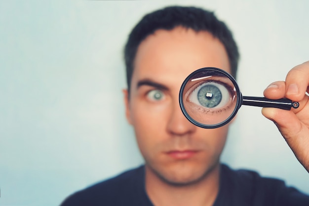 Potrait van jonge man kijken door vergrootglas op witte onscherpe achtergrond. Mening aan mannelijk blauw oog door de lens. Loep met macrooog in de hand van de persoon op de voorgrond. Informatie zoeken.