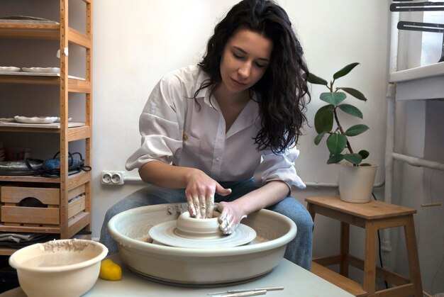 白いシャツとブルージーンズを着た陶芸家が速く回転する陶芸者の車輪で鉢や花瓶を作ります