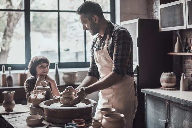 Pottering together is fun. Cheerful young man and little boy making ceramic pot on the pottery class