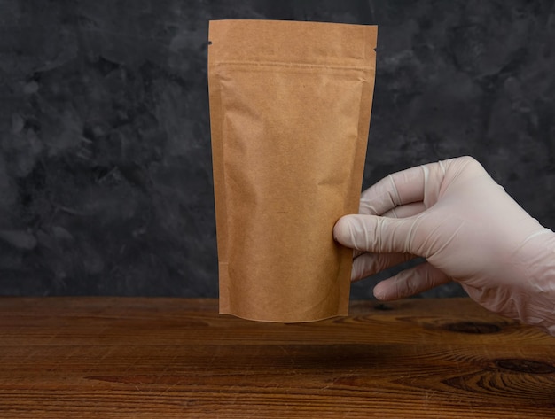 Pouch bag mockup Hand in protective gloves holds brown kraft paper pack with coffee beans wooden background