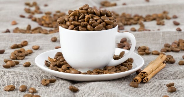 Poured in a white mug roasted very tasty coffee grains on a tablecloth, next to it are sticks of fragrant and pleasant cinnamon