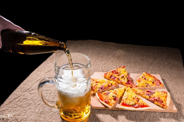 Pouring Bottled Beer into Glass Mug with Meal of Fresh Baked Pizza Arranged in Slices on Wooden Cutting Board on Burlap Covered Table